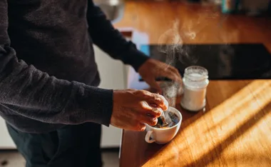 Person stirring steaming coffee in a cup beside a jar on a wooden countertop.