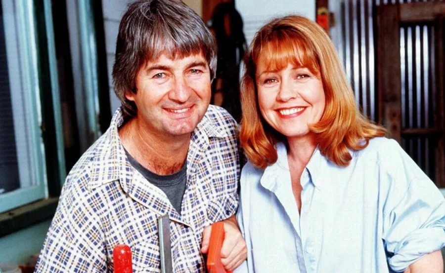 A man in a plaid shirt and a woman in a light blue blouse smile warmly, standing side by side indoors.