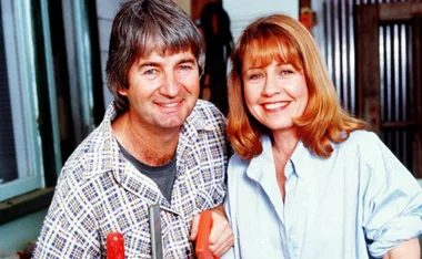 A man in a plaid shirt and a woman in a light blue blouse smile warmly, standing side by side indoors.