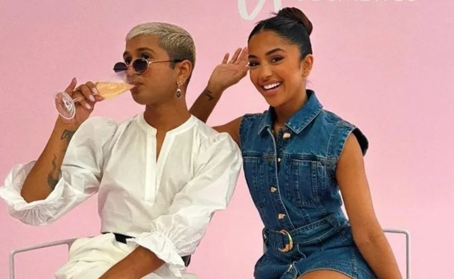 Two people smiling; one drinks champagne, the other waves, seated against a pink background.