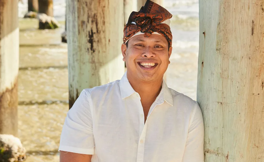 Man in a white shirt and traditional headwear smiling under pier.