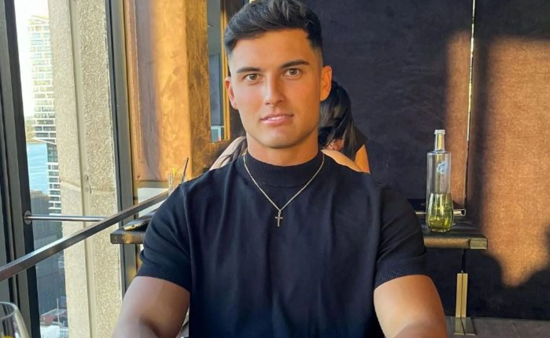 A person with short dark hair, wearing a black shirt and a cross necklace, sitting in a restaurant with a city view.