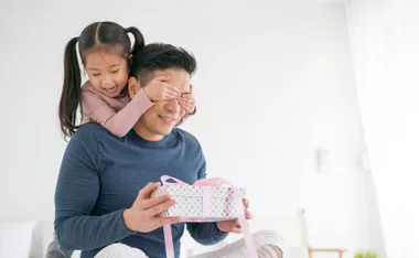 Father receiving a gift with daughter covering his eyes, smiling in a bright room.