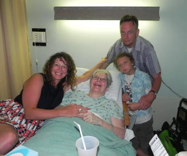 Family gathered around an elderly woman in a hospital bed, smiling and expressing warmth and togetherness.