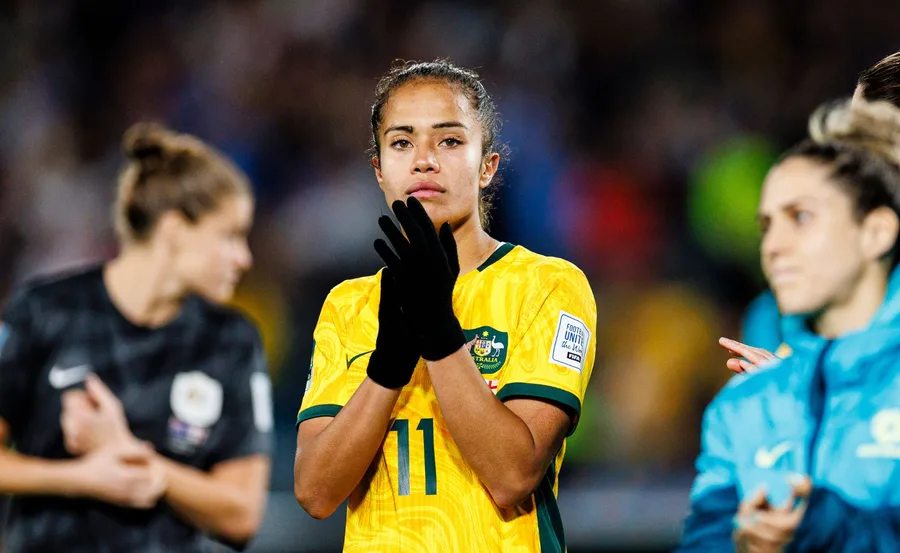 Australian soccer player in yellow jersey clapping, wearing black gloves, with fellow players in blurred background.