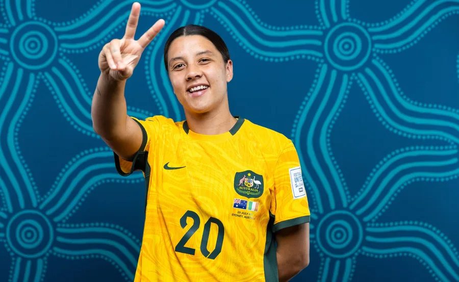 A soccer player in a yellow jersey with "20" displays a peace sign, smiling in front of a patterned blue backdrop.