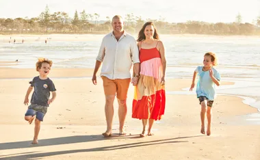 Family walking on the beach, parents holding hands and children running beside them in the sunshine.