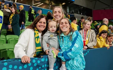 A group of people, including a young child, posing and smiling in a stadium with green seats and spectators in the background.