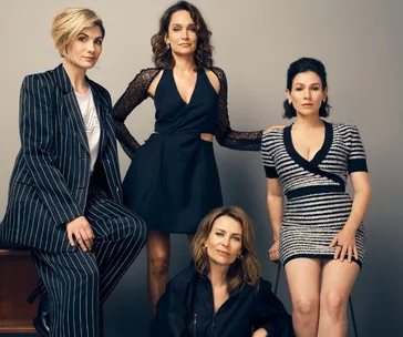 Group of four women posing together; three standing and one seated, dressed in stylish outfits.