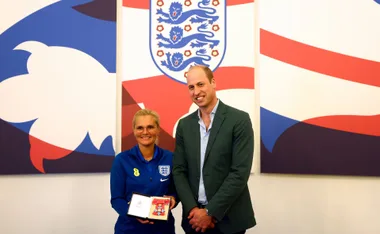 Two people smiling, one holding an award, standing in front of an England football emblem background.