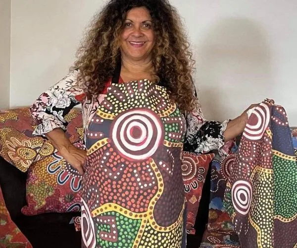 An artist wearing a colorful apron with abstract indigenous patterns stands smiling, displaying a matching fabric piece.