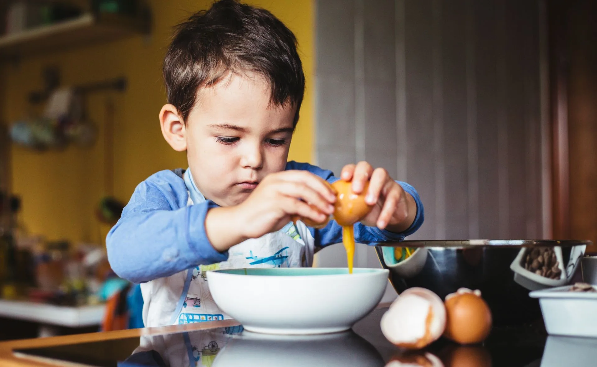 Young child cracking an egg into a bowl in a kitchen setting.