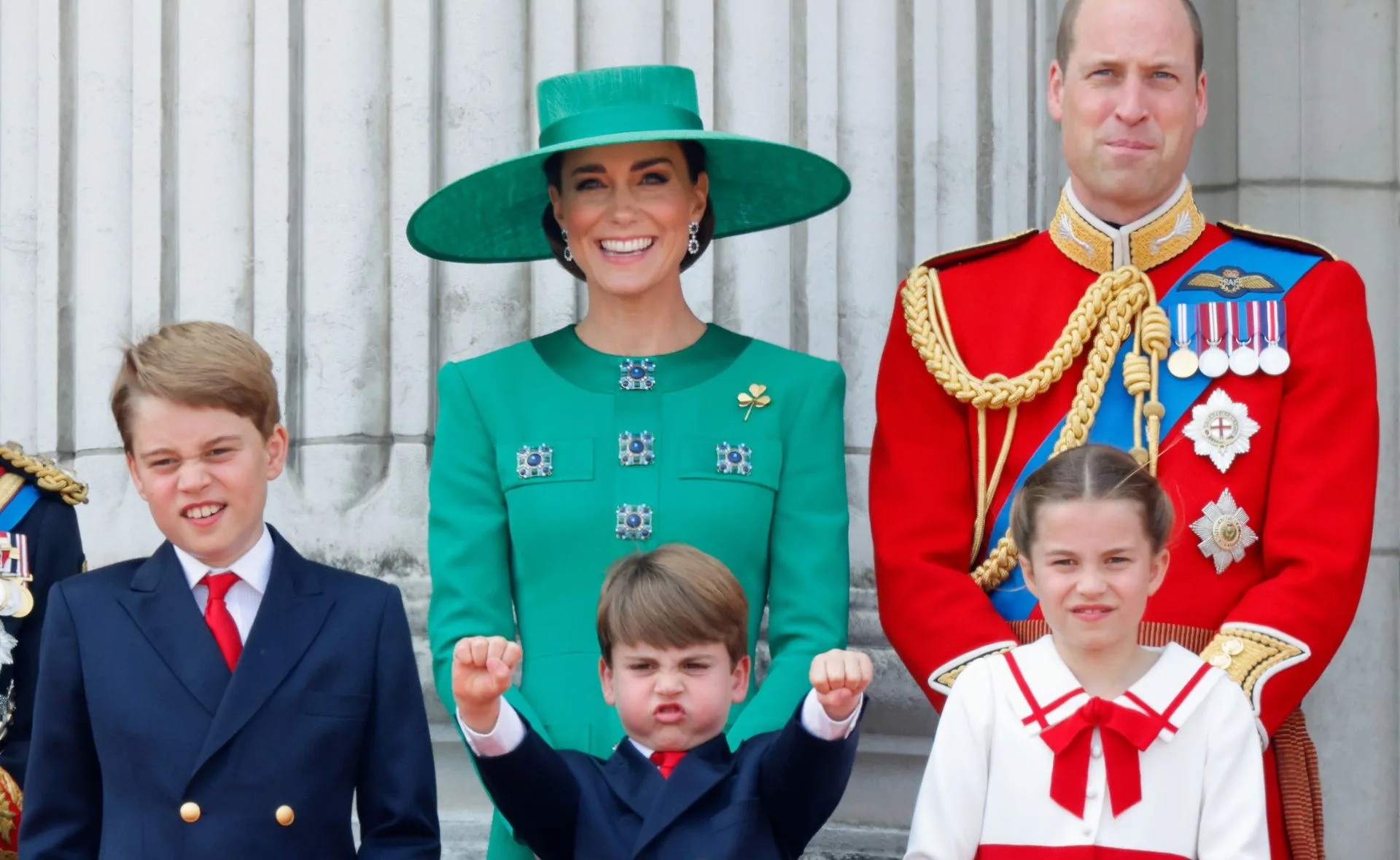 Royal family members stand together, smiling, with young child playfully posing in front.