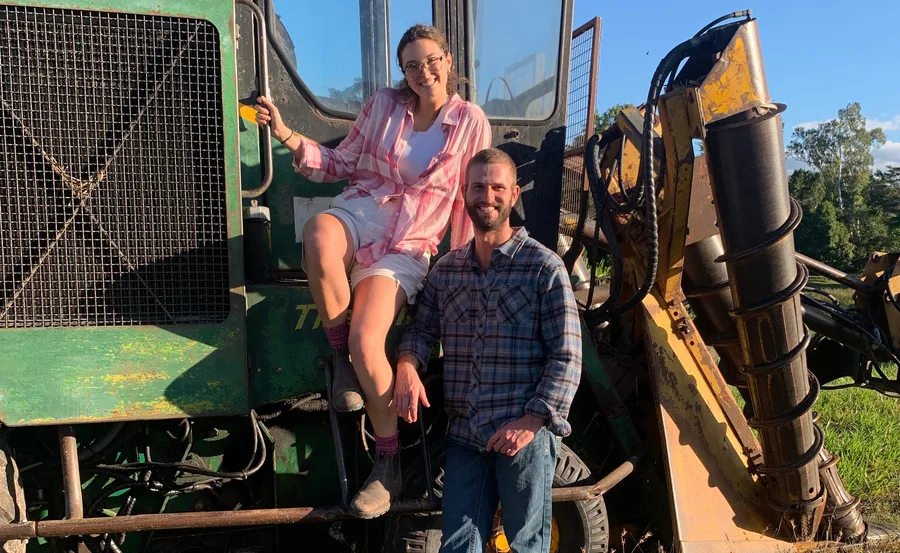 A woman in a pink shirt sits on a green tractor beside a smiling man in a plaid shirt, outdoors in sunny weather.