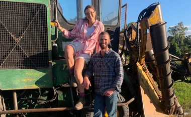A woman in a pink shirt sits on a green tractor beside a smiling man in a plaid shirt, outdoors in sunny weather.