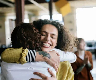 Woman with curly hair smiling widely while hugging another person; joyful indoor setting.