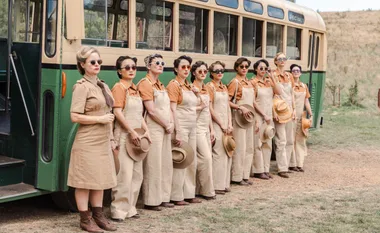 Women in 1940s attire stand beside a vintage bus, wearing tan uniforms and holding hats, reminiscent of WWII era workers.