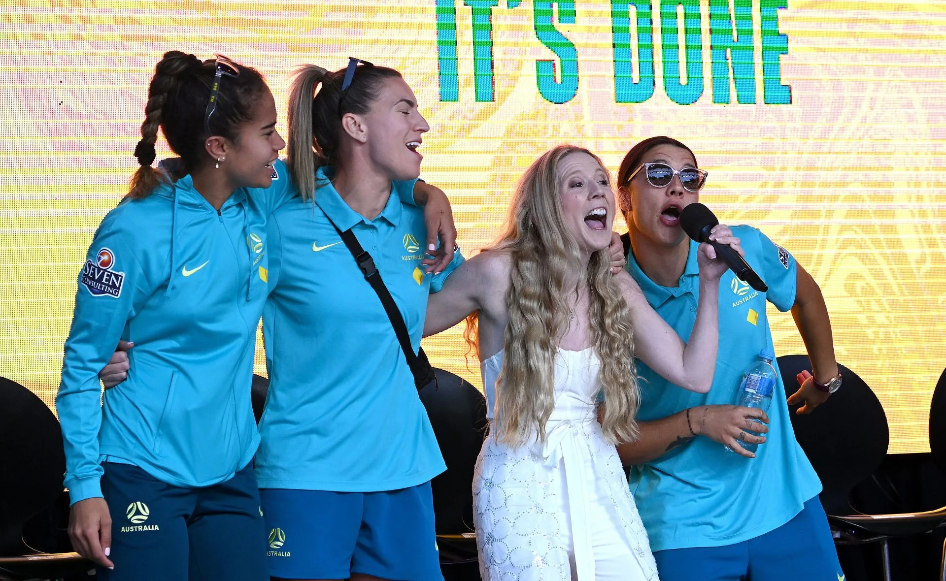 A woman in white sings with three sportswomen in blue jerseys on stage, smiling and holding a microphone.
