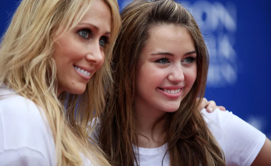 Two women with long hair smiling at a public event, standing closely with a blue background.