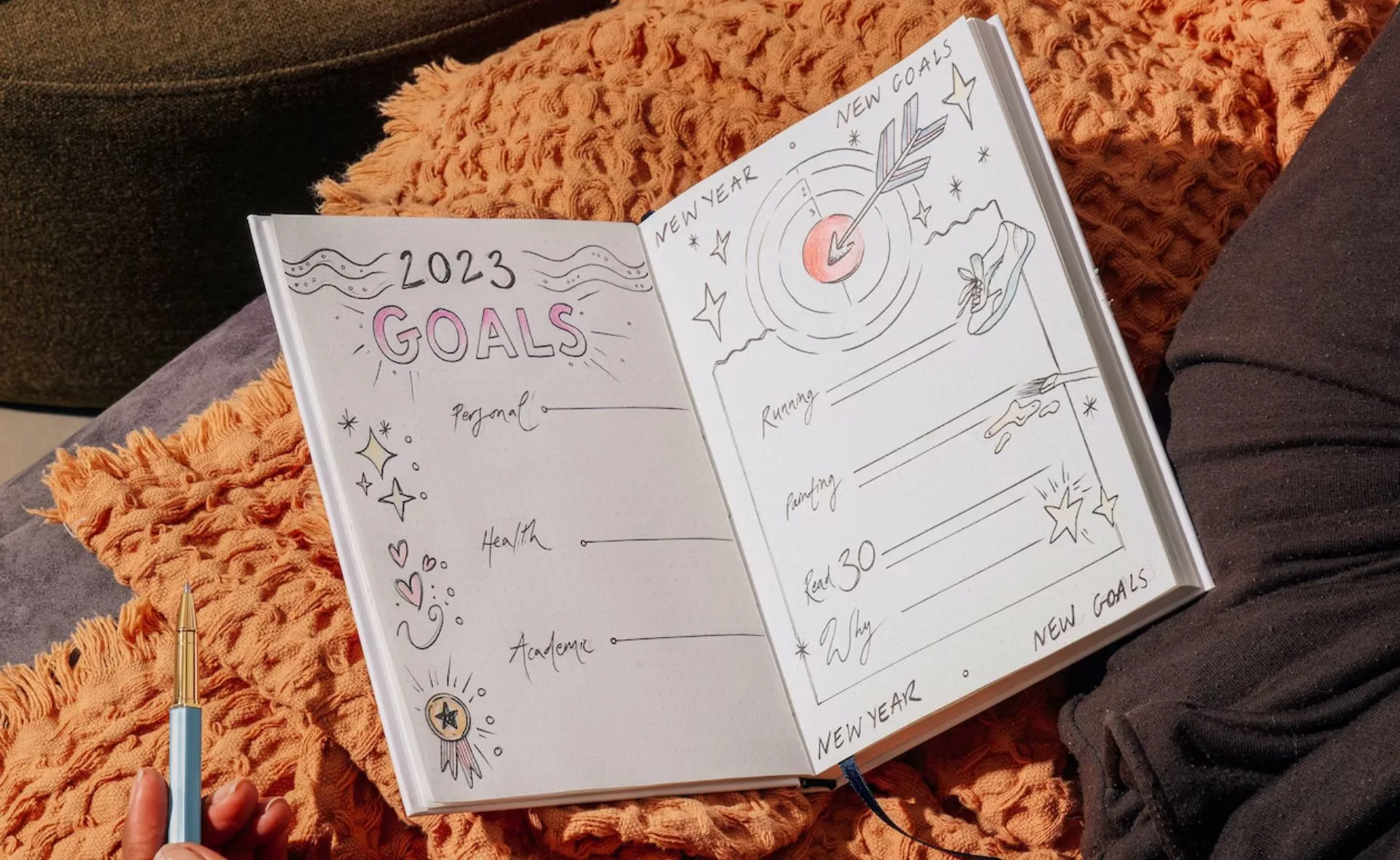 Open bullet journal with "2023 Goals" and illustrations, next to an orange cushion and a person holding a pen.