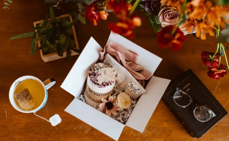 Candle subscription box with a candle, tea, flowers, glasses, and a book on a wooden table.