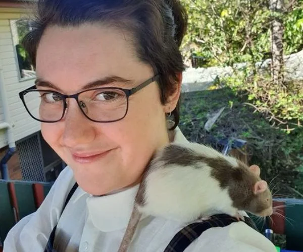 Portrait of a smiling person with glasses, outdoors, with a white and brown rat on their shoulder.