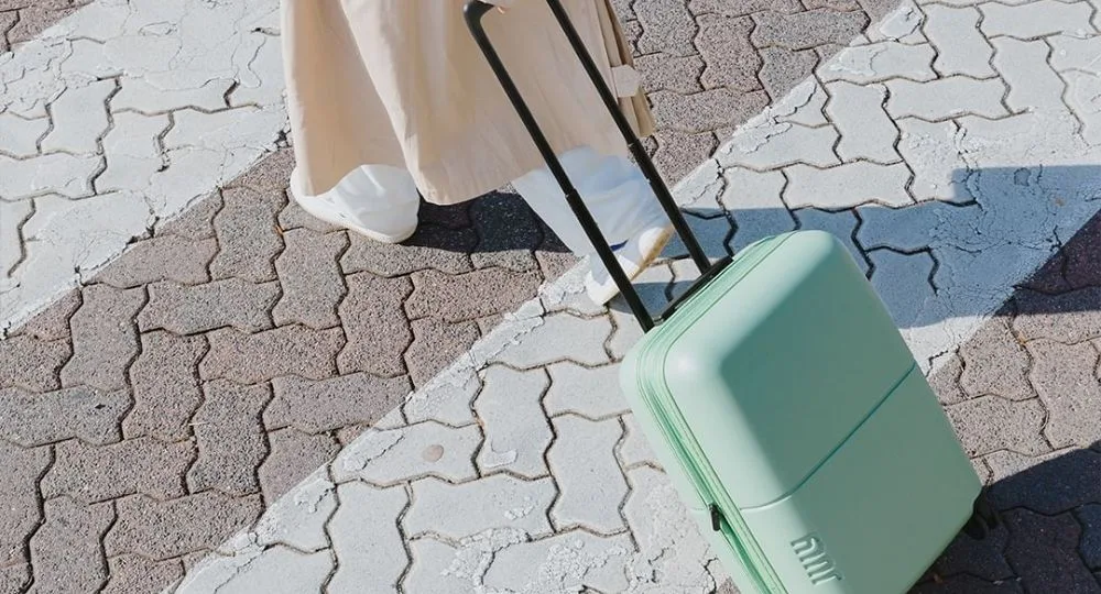 Person in beige coat pulling mint green rolling luggage on paved path.