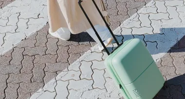 Person in beige coat pulling mint green rolling luggage on paved path.