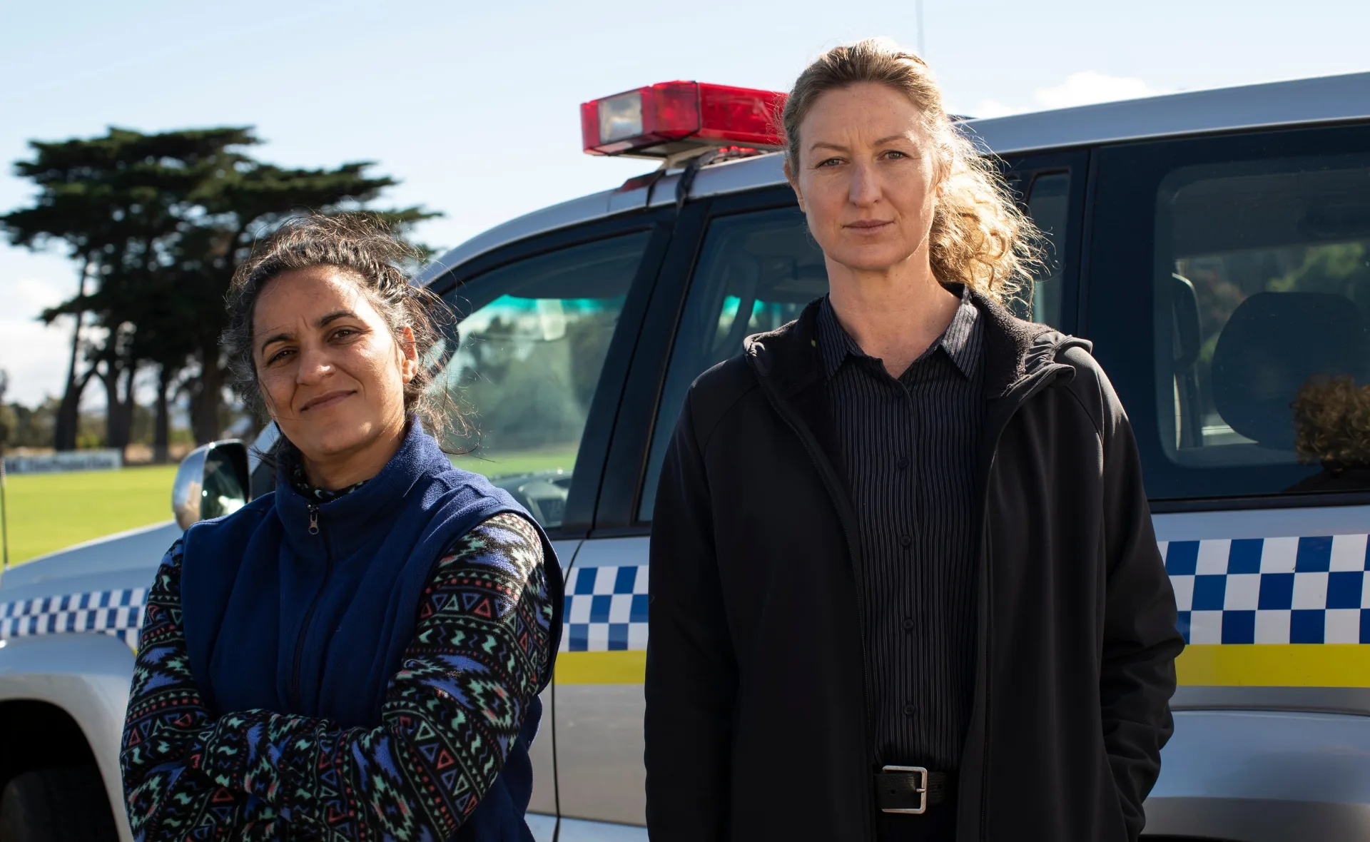 Two women stand near a police vehicle in a sunny outdoor setting.