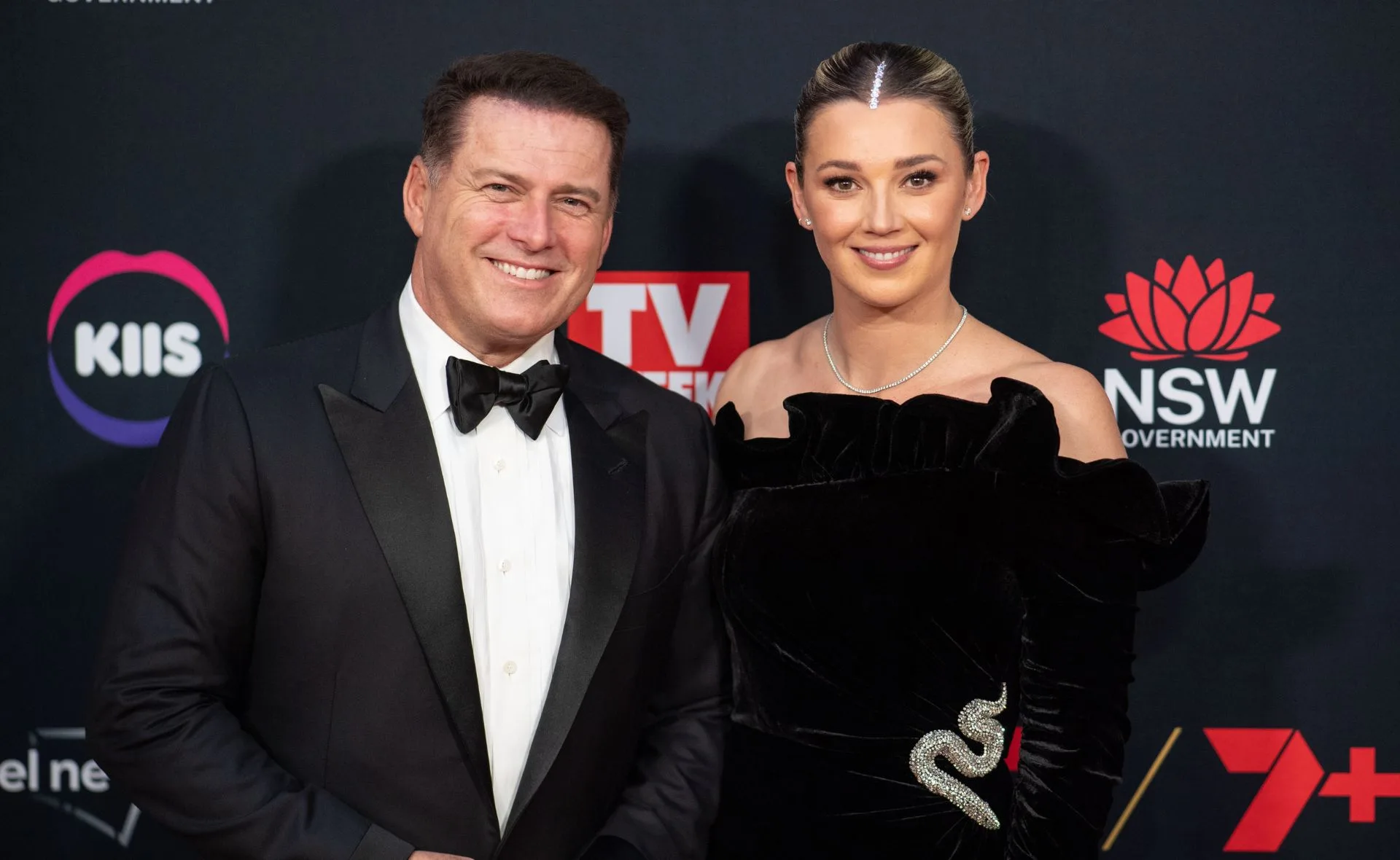 A couple in formal attire smiling on the red carpet, with event logos in the background.