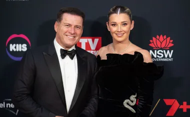A couple in formal attire smiling on the red carpet, with event logos in the background.