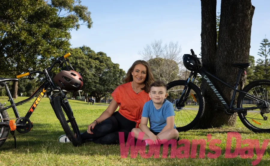 Woman and boy sit on grass with bicycles and trees around, smiling in a park setting.