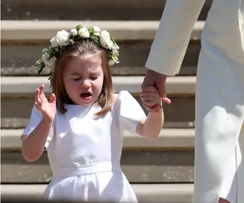 A young girl in a white dress and floral crown sneezes while holding an adult's hand, walking down steps.