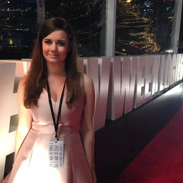 A woman in a light pink dress on a red carpet with a lanyard, standing in front of large event signage.