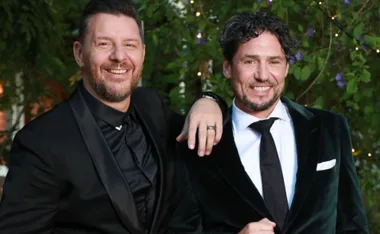 Two men in formal suits smiling, standing in an outdoor setting with greenery in the background.