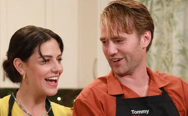 A woman and a man smiling in a kitchen, wearing aprons.
