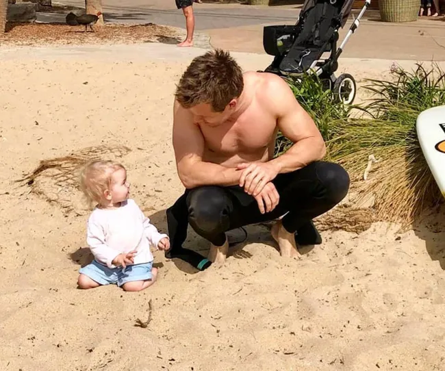 Shirtless man crouches on sand, interacting with a seated toddler. A stroller and surfboard are nearby.