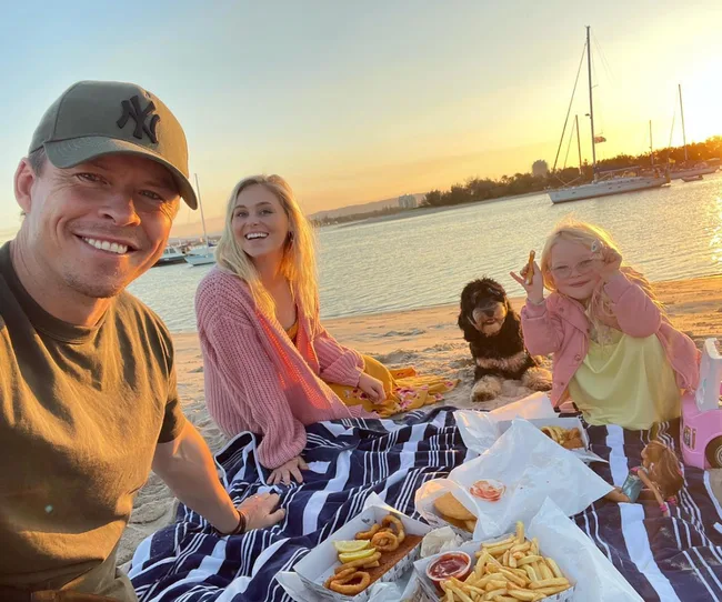 Family enjoying a picnic on the beach during sunset with food and a dog sitting nearby.