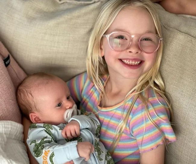 A smiling young girl in glasses holds a baby with a pacifier, sitting on a couch with a cozy blanket nearby.