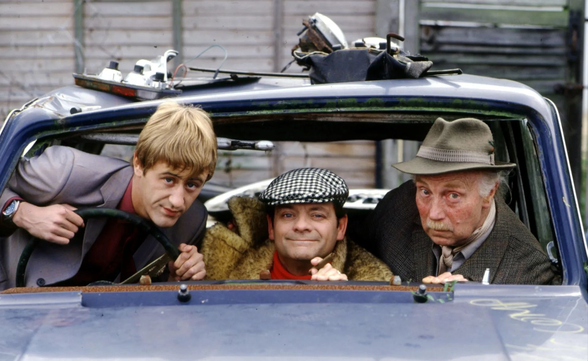 Three men sitting in a beat-up car, one at the wheel, one in the middle, and the other on the right wearing a hat.