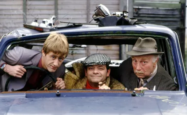 Three men sitting in a beat-up car, one at the wheel, one in the middle, and the other on the right wearing a hat.