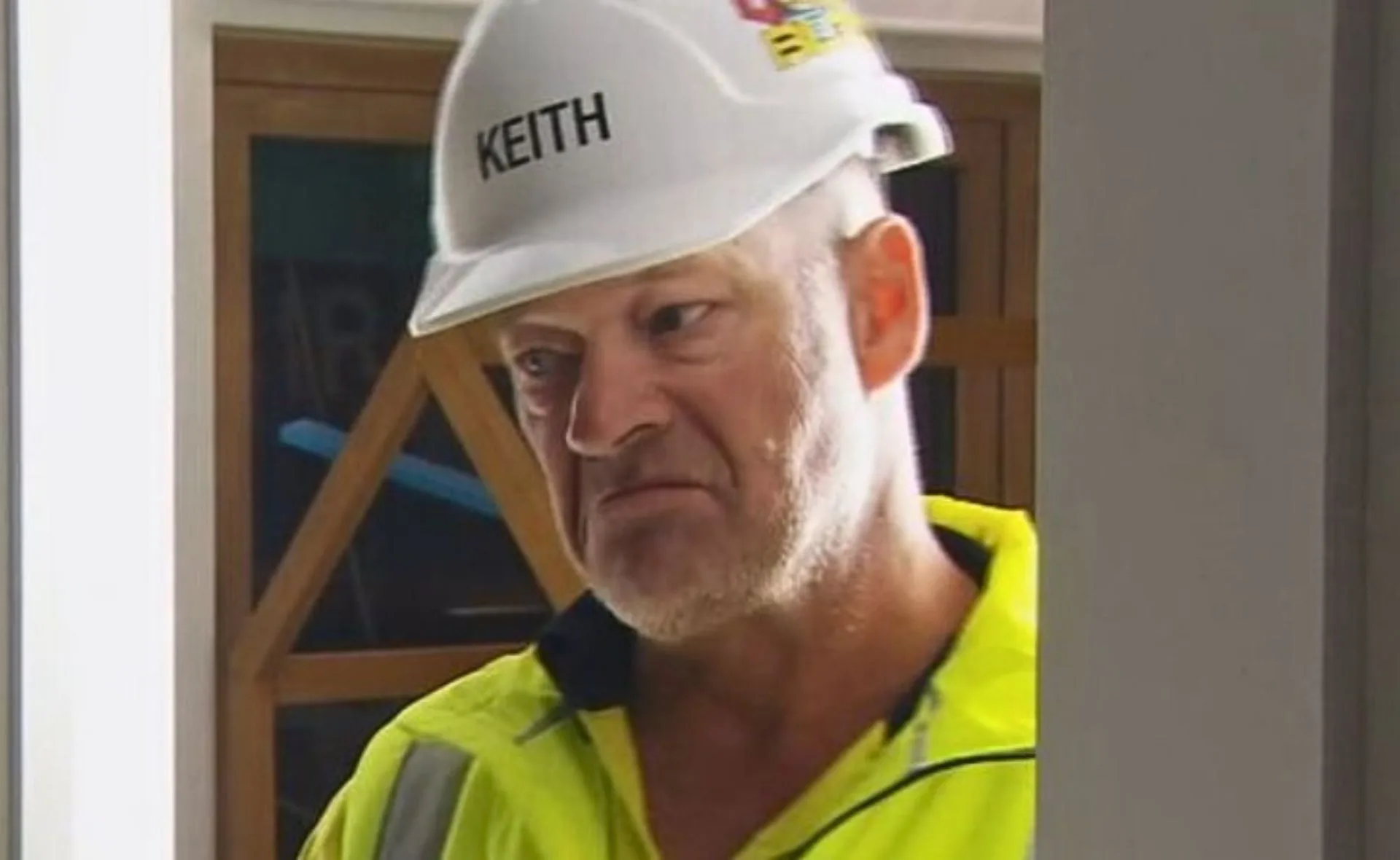 Man in construction gear and helmet labeled "Keith" with a serious expression indoors.