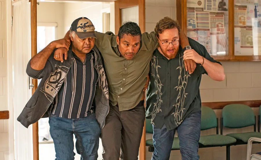 Three men support each other as they walk down a hallway in a building, with chairs visible in the background.