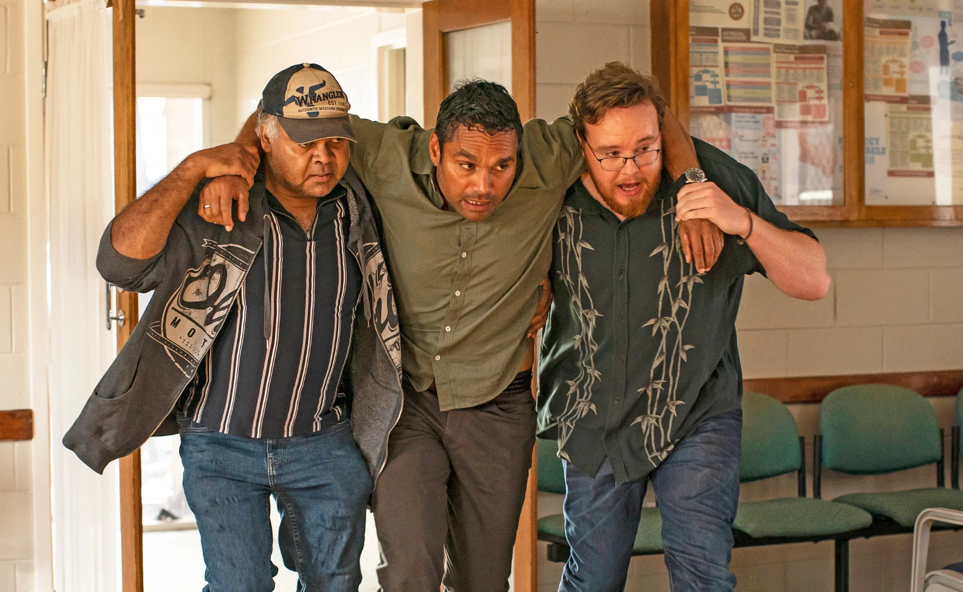 Three men support each other as they walk down a hallway in a building, with chairs visible in the background.