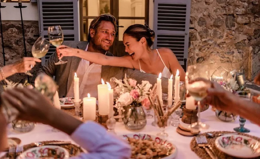 A couple smiling, toasting with wine glasses at a candlelit table with flowers and plates, surrounded by friends.