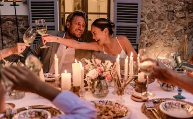 A couple smiling, toasting with wine glasses at a candlelit table with flowers and plates, surrounded by friends.