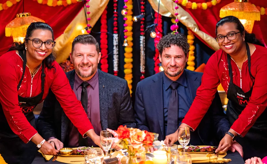 Four people at a festive dinner, two standing in aprons serving food to seated guests, with colorful decorations.