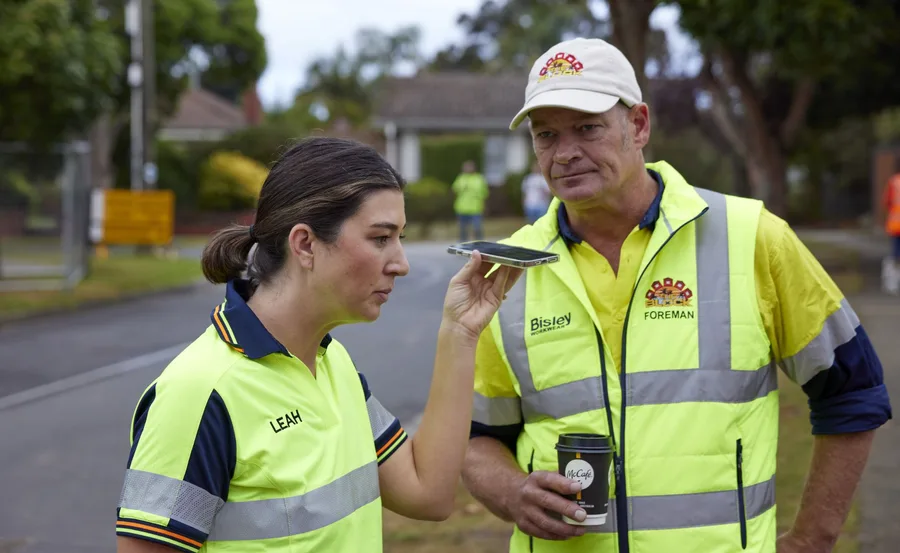 Two construction workers in high-visibility vests; one is speaking into a phone, the other holds a coffee cup.