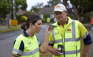 Two construction workers in high-visibility vests; one is speaking into a phone, the other holds a coffee cup.