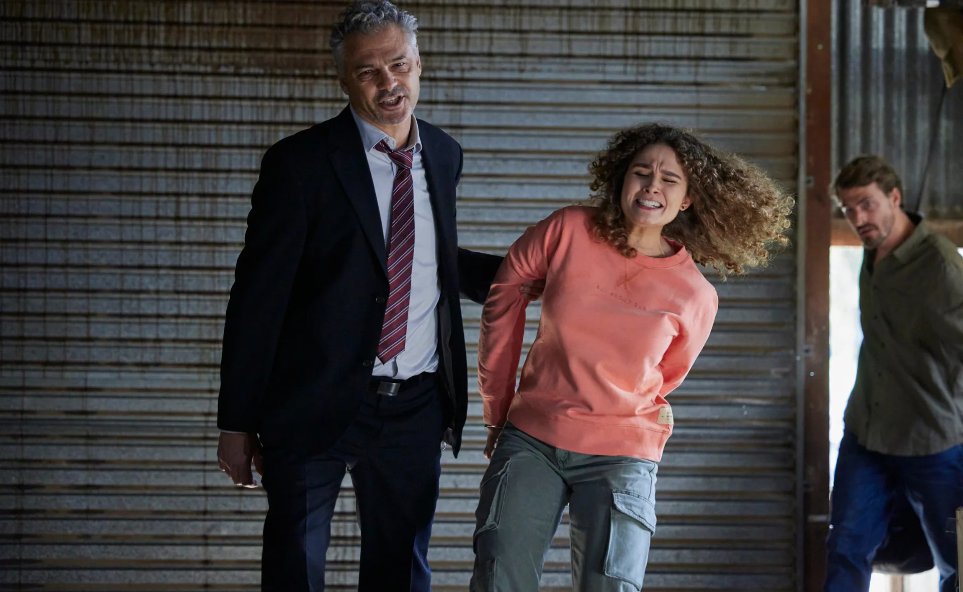 A man in a suit holds a woman in a pink sweater, who looks distressed, with another man in the background.